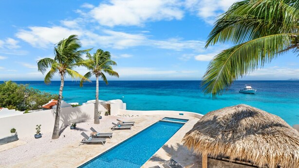 A pool area with lounge chairs, a thatched-roof structure, and palm trees overlooking a clear blue sea. - Kas di Bon Beis - Spacious Oceanfront Villa with Full A/C (Kralendijk)