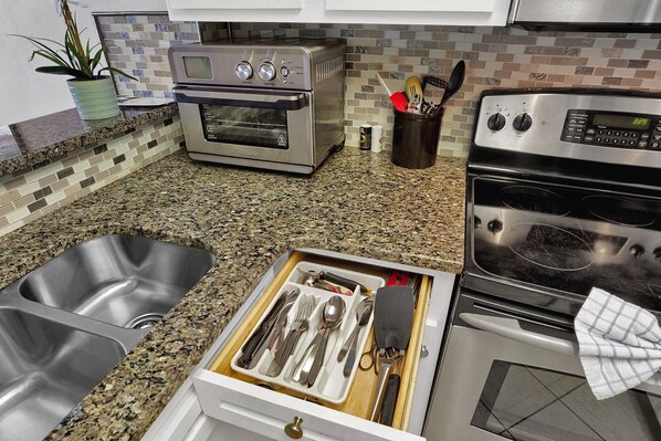 A kitchen with granite countertops, stainless steel appliances, and a drawer organizer with utensils. - Beach Colony Resort - 714 (Myrtle Beach)