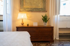 A hotel room with a bed, a wooden nightstand with a lamp, a potted plant, and a painting on the wall.