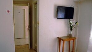 Egy szállodai szoba, amelyben található egy ágy, a falra szerelt televízió, egy kis, fából készült asztal, rajta egy virágvázával, és egy ajtó, amely egy másik szobába nyílik.
