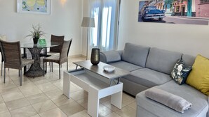 A modern living room with a grey sofa, a white coffee table, wicker chairs, a dining table with a potted plant, and a cityscape painting on the wall. - Villa Horizon Salobre 7 (San Bartolomé de Tirajana)