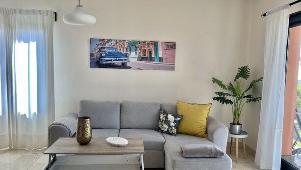 A living room with a grey sofa, a small table with a vase, a plant, and a painting of a street scene. - Villa Horizon Salobre 7 (San Bartolomé de Tirajana)