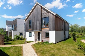 Une maison moderne à deux étages, dotée d’un toit en pente, de grandes fenêtres et d’un espace extérieur aménagé, avec un chemin en gravier.