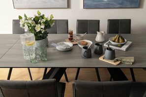 Une table à manger est dressée avec divers objets, notamment un vase de fleurs, une carafe, un saladier rempli de poires et un livre intitulé « L’anatomie des cabanes dans les arbres ».