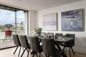 Un espace repas moderne avec vue sur un plan d’eau, un barbecue à l’extérieur et deux tableaux abstraits accrochés au mur.