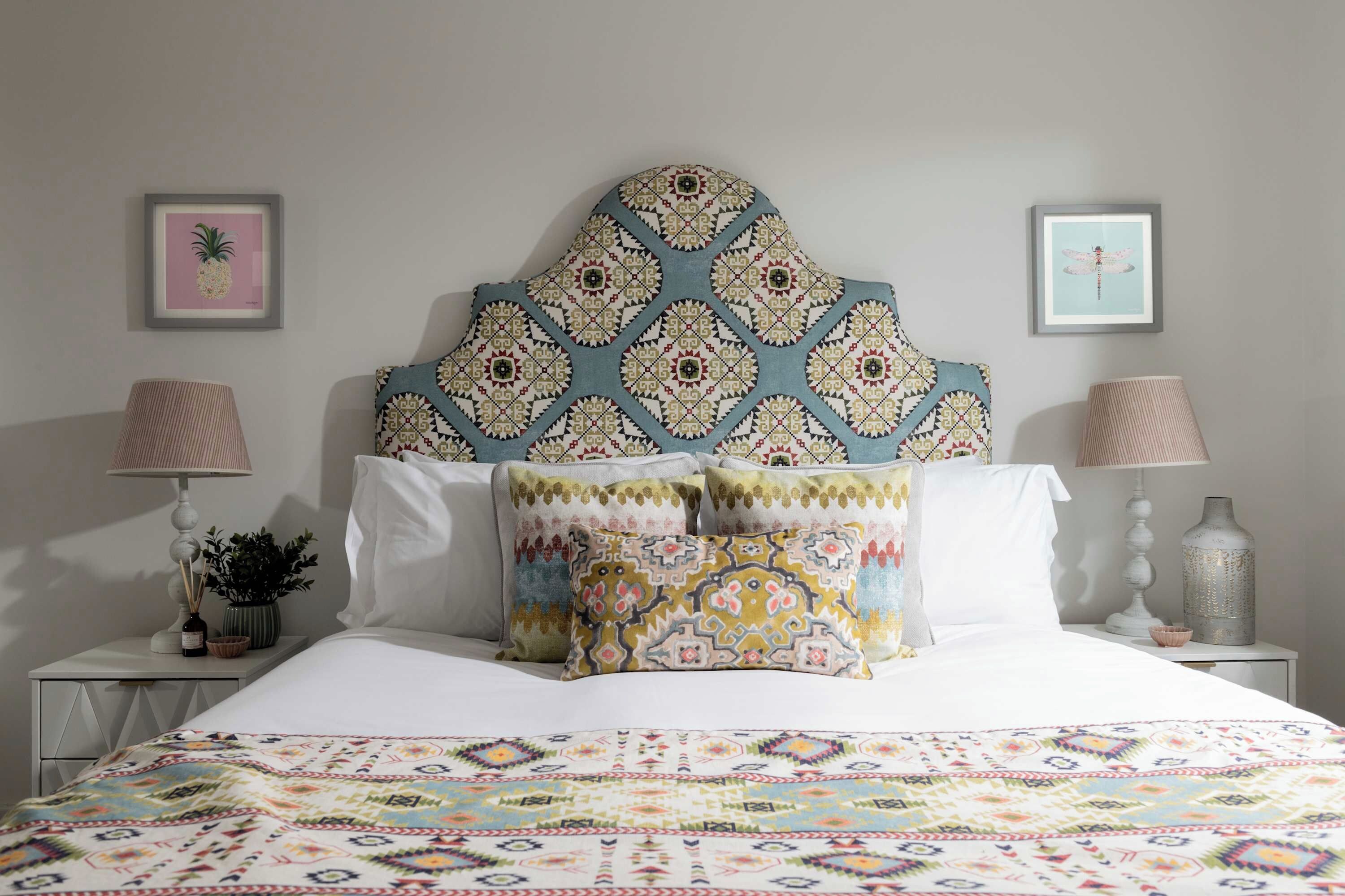 Ein ordentlich bezogenes Bett mit gemusterten Kissen und einer farbenfrohen Steude, ein Kopfteil mit geometrischem Muster und zwei gerahmte Bilder an der Wand.
