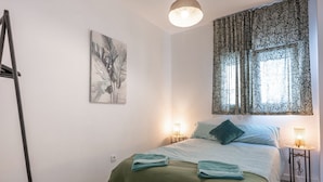 A bedroom with a bed, two bedside lamps, a green blanket, a painting on the wall, and a window with patterned curtains. - Océano: Acogedor y Luminoso Apartamento (Santa Cruz de Tenerife)