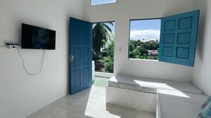A room with a TV mounted on the wall, two open doors leading to a balcony, and a bed with a white mattress. - Casas Astro Rei (Cairu)