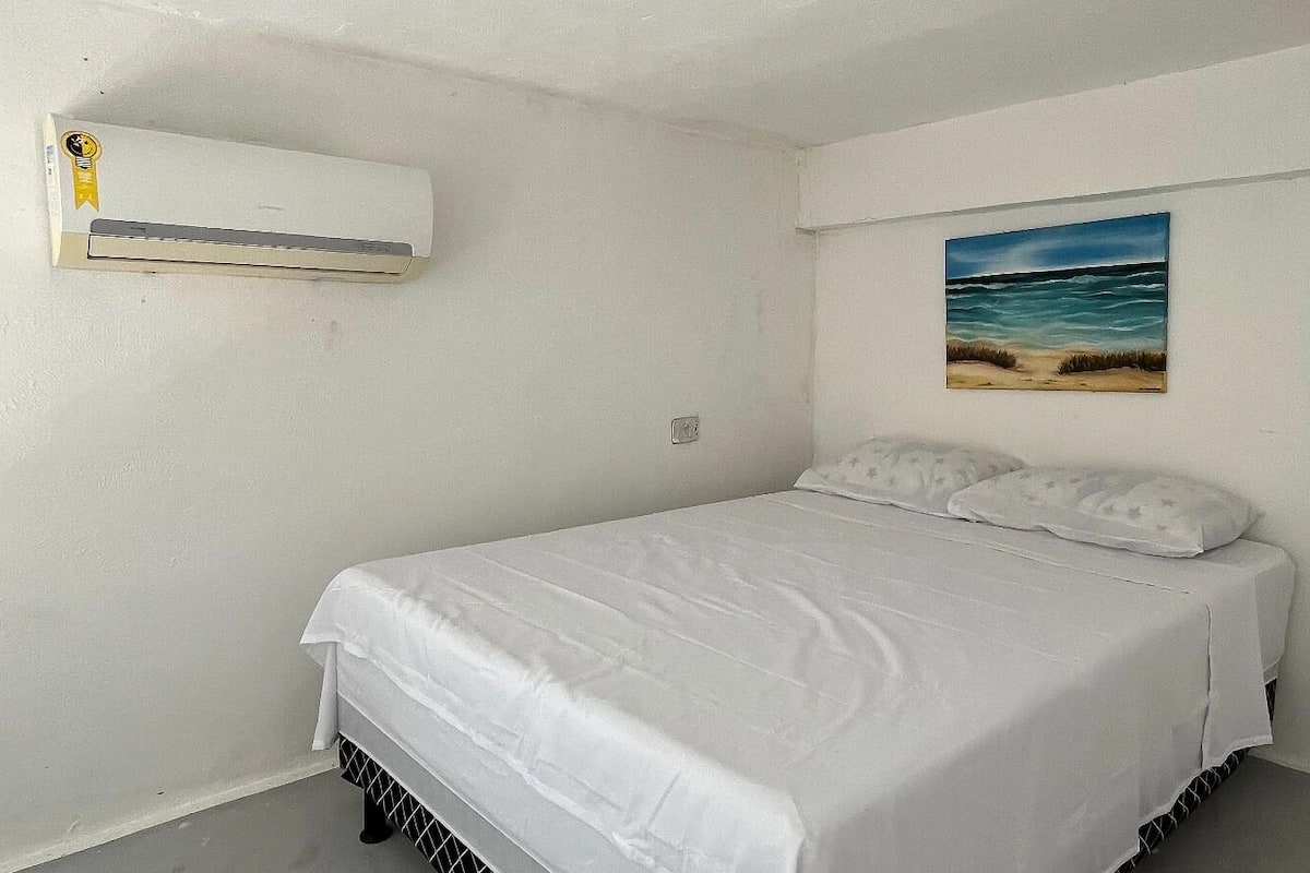 A bedroom with a bed, white walls, a painting of a beach, and an air conditioning unit.