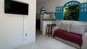 A compact living space with a flat-screen TV mounted on the wall, a kitchenette with a fridge, and a sofa with a red-striped throw blanket. - Casas Astro Rei (Cairu)