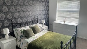 A bedroom with a patterned wallpaper, a bed with green bedding, a nightstand with a lamp, a dresser, and a window with blinds.