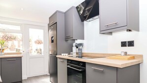 A modern kitchen with grey cabinets, a built-in oven, and a window with a view of a garden. - Lilly's Cottage (Dawlish)