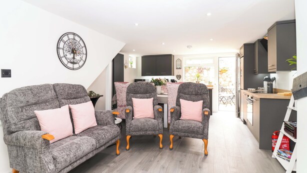 A modern living room with a grey sofa and matching armchairs, a dining area with a pink tablecloth, and a kitchen with dark cabinets. - Lilly's Cottage (Dawlish)