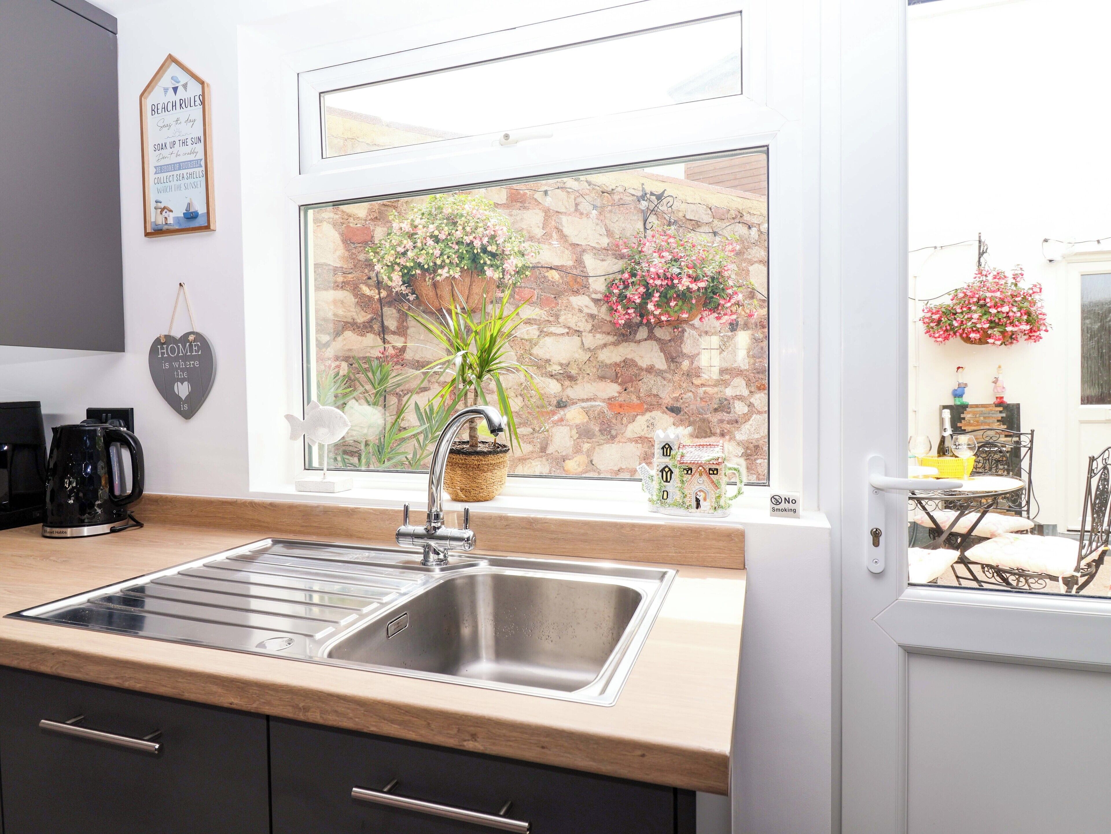 A kitchen with a double sink, a coffee maker, and a window with a view of a stone wall and hanging flower baskets.