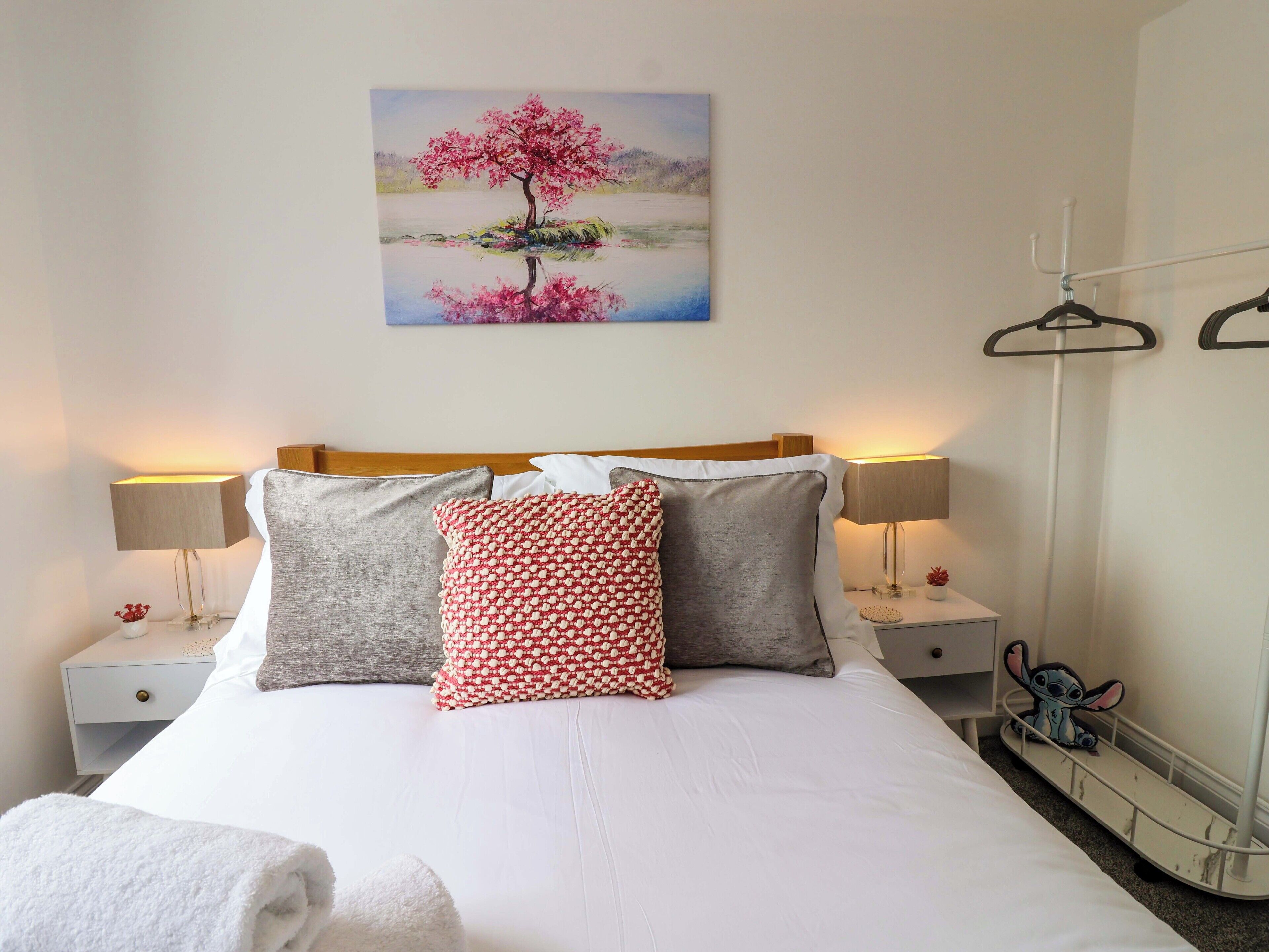 A neatly made bed with white bedding, two bedside lamps, a wall-mounted painting of a tree, and a small bedside table with a lamp.