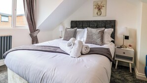 A neatly made bed with a grey headboard, white bedding, and two rolled towels. There is a bedside table with a lamp and a small plant. A window with curtains is visible, and a painting hangs on the wall. - Lilly's Cottage (Dawlish)