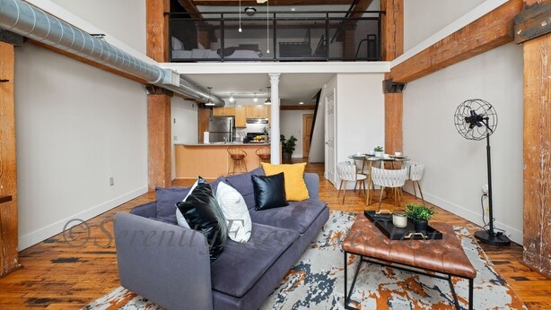 Un loft spacieux avec un plafond haut, des poutres apparentes, un canapé gris avec des coussins, une table basse et un coin repas avec des chaises et une petite table.