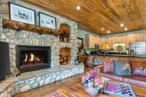 A stone fireplace with a fire, a wooden ceiling, a kitchen area, and a leather sofa.