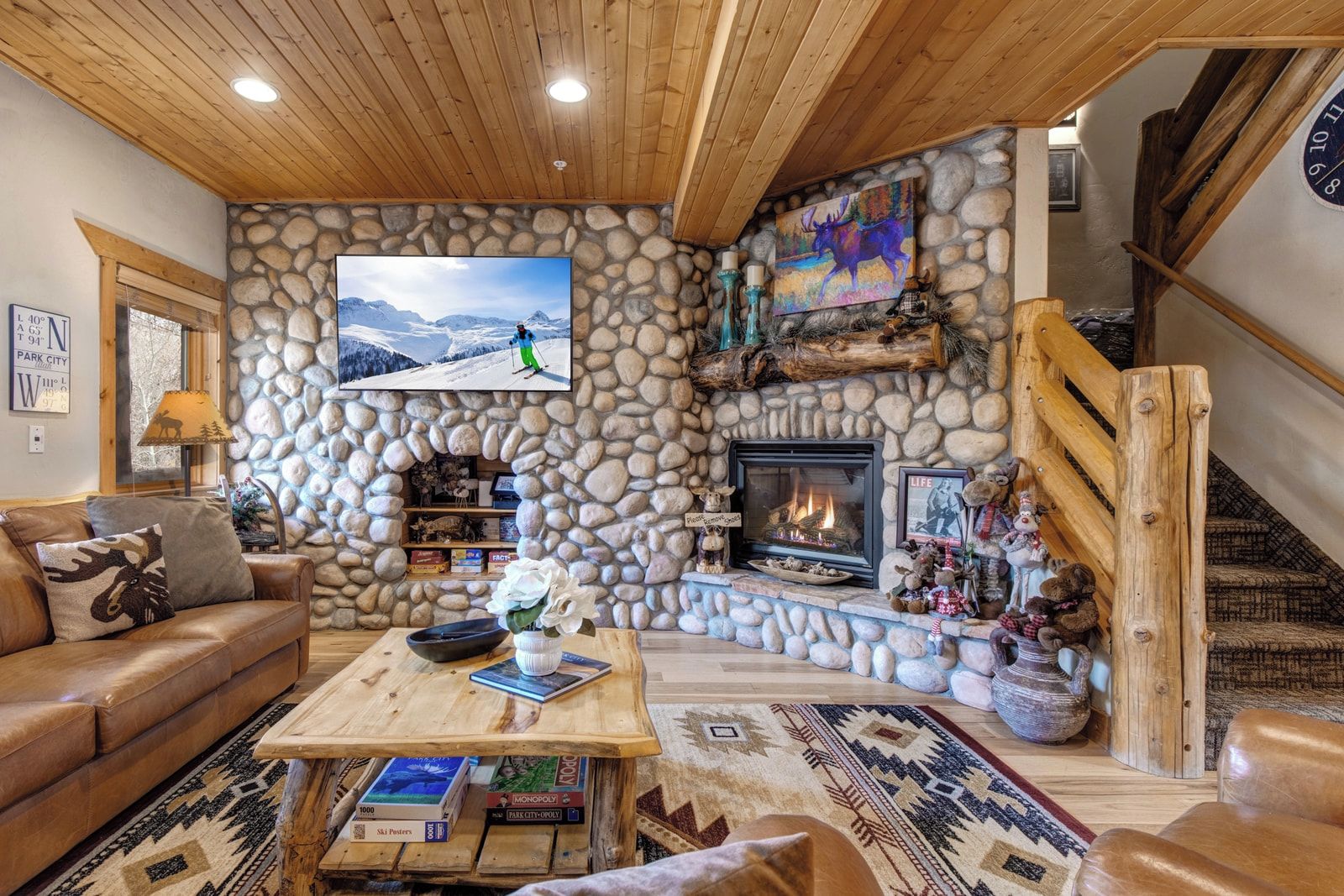 A cozy living room with a stone fireplace, a television mounted on the wall, and a wooden staircase.