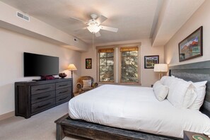 Une chambre à coucher comprenant un lit, un téléviseur, une commode, une chaise, un ventilateur de plafond et une fenêtre donnant sur des arbres.