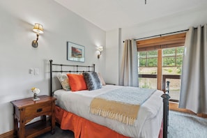 Une chambre à coucher avec un lit, des lampes de chevet, une table de nuit, une fenêtre avec des rideaux et une vue sur la verdure à l’extérieur.