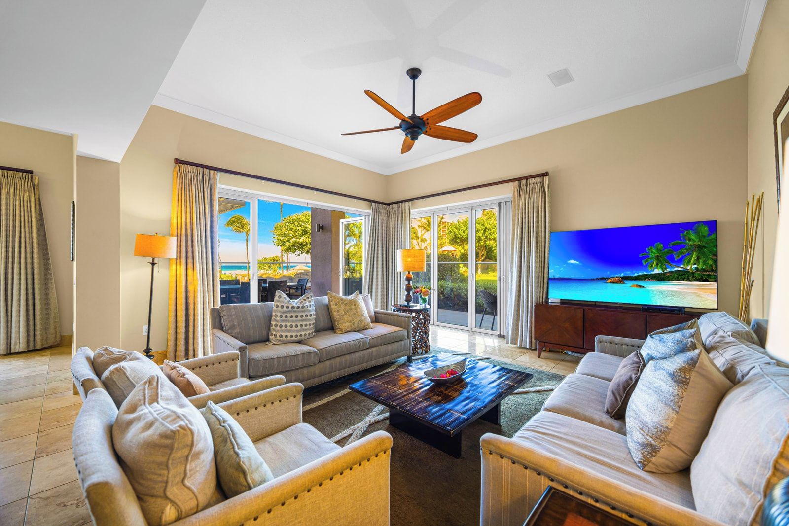 A spacious living room with a ceiling fan, large windows, a flat-screen TV, and a view of the beach.
