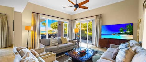 A spacious living room with a ceiling fan, large windows, a flat-screen TV, and a view of the beach.