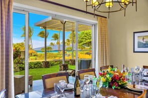 A dining area with a table set for a meal, large windows offering a view of a landscaped garden and a beach. - Honua Kai | Ocean View 4 BR, Sleeps 14 | Car Incl. w/ 6+ Nights | HKH ML-1351 by KBM (Lahaina)