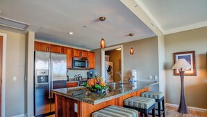 A kitchen with wooden cabinets, a stainless steel refrigerator, a microwave, and a sink. There are three striped bar stools and a decorative plant on the island.
