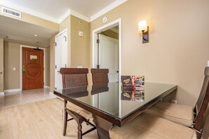 Une chambre d’hôtel avec une table à manger en verre, deux chaises marron à revêtement en tissu, un luminaire fixé au mur et une affiche accrochée au mur.