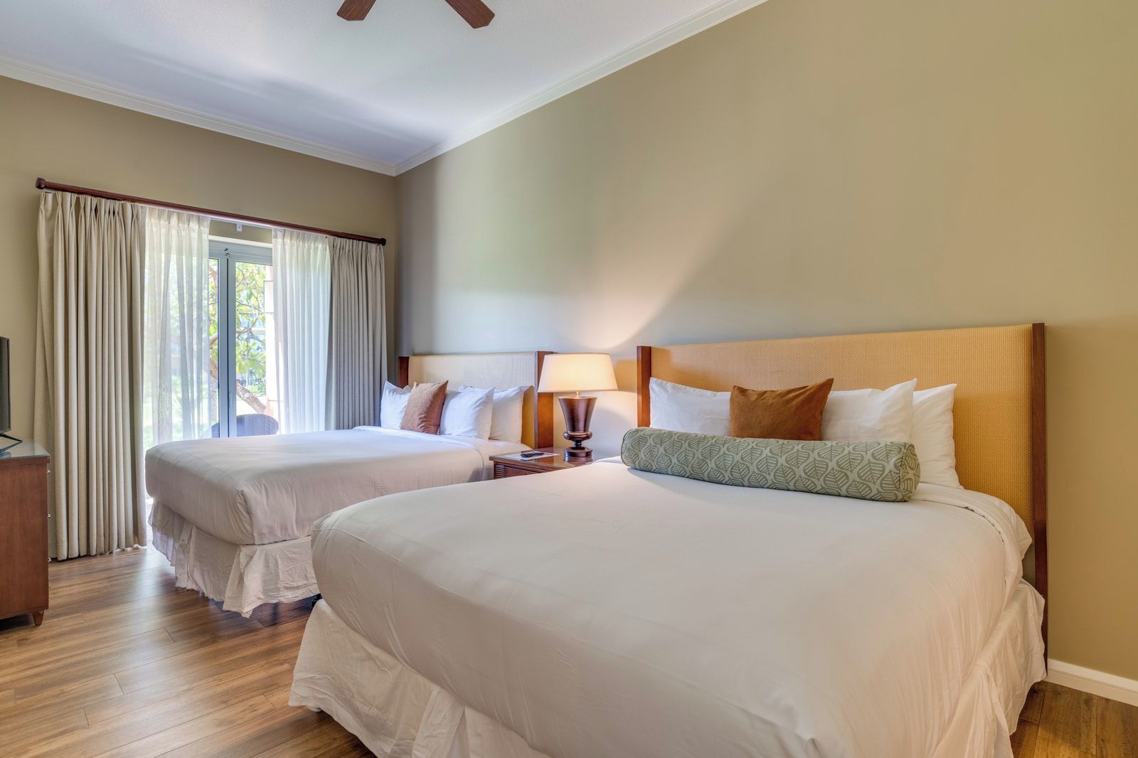 A hotel room with two beds, a wooden headboard, a nightstand with a lamp, and a window with curtains.