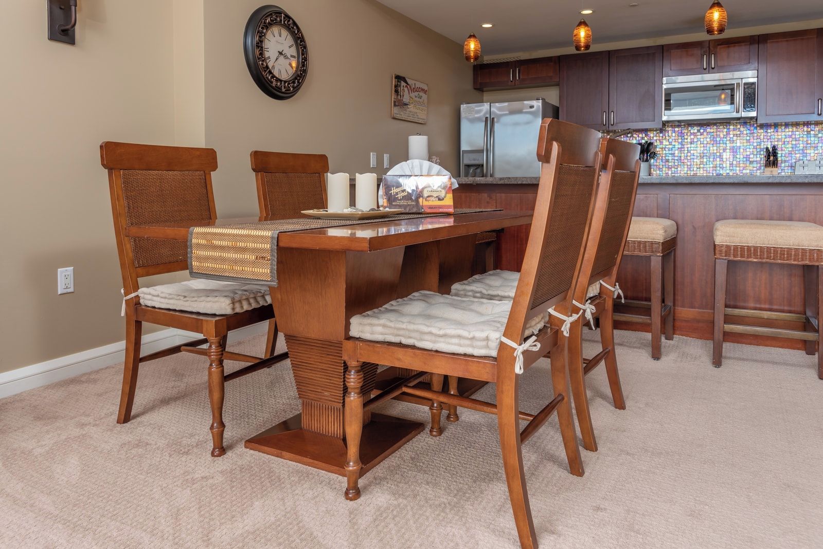 Een eethoek met een houten tafel en stoelen, een keuken met donkere kastjes en een betegelde achterwand, en een wandklok.