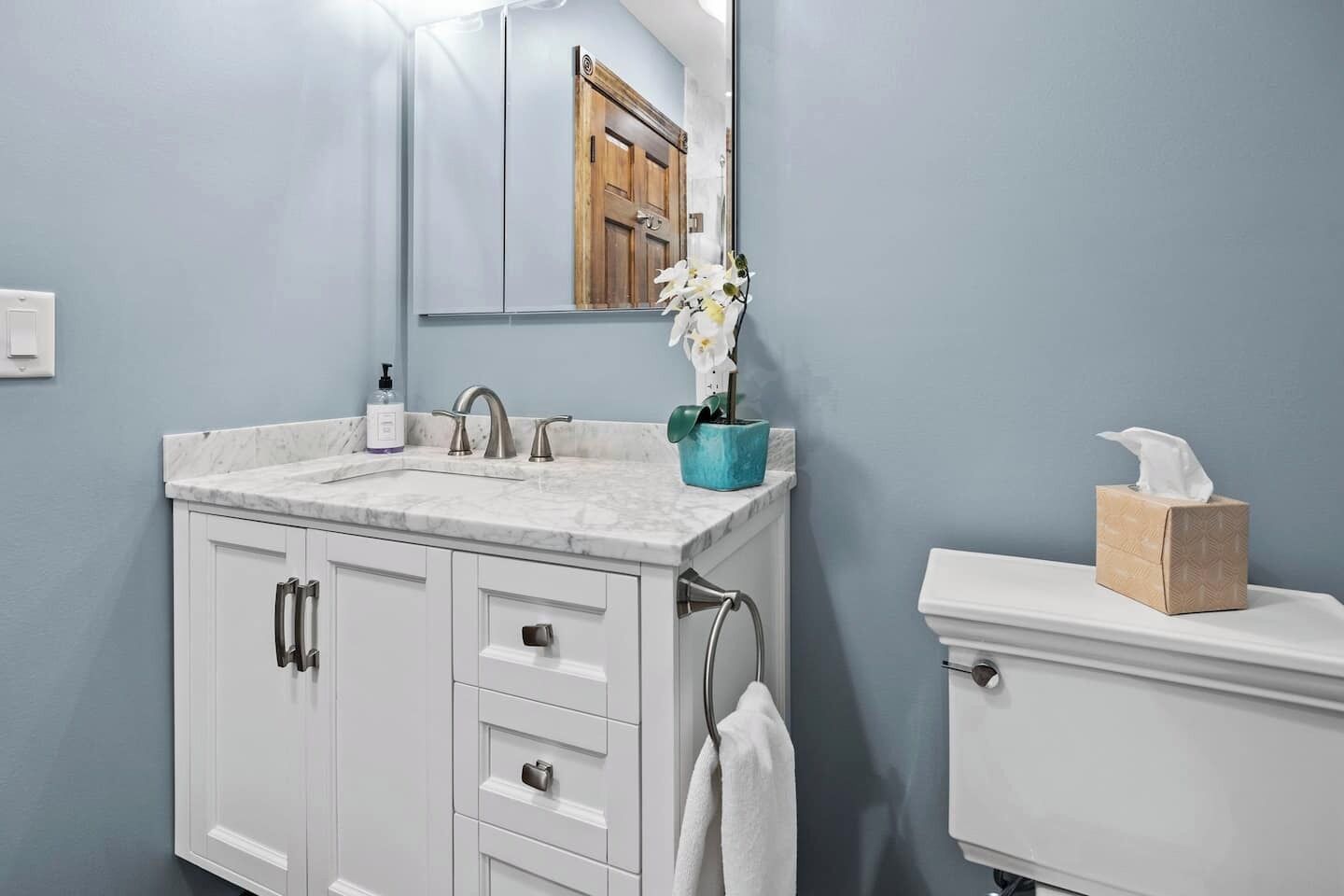 A bathroom with a marble sink, white cabinets, a mirror, a toilet, and a towel hanging on the wall.