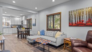 Una sala de estar moderna con un sofá gris, una mesa de centro de madera y un sillón de cuero marrón. Se ve un área de comedor con una mesa y sillas de madera al fondo.
