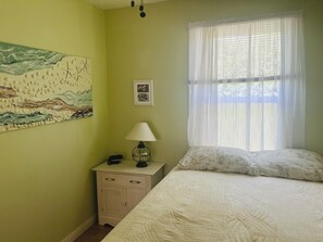 A bedroom with a bed, a nightstand with a lamp, a wall painting, and a window with sheer curtains. - 2 bdm cottage in the BLUFF, ONLY 3 min walk to PARLEE. AC, WiFi and 2 sunrooms (Shediac)