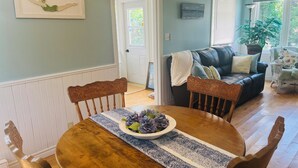 Un coin repas avec une table en bois, des chaises et un vase rempli de fleurs violettes. On aperçoit, à l'arrière-plan, un canapé en cuir noir et un fauteuil en osier.