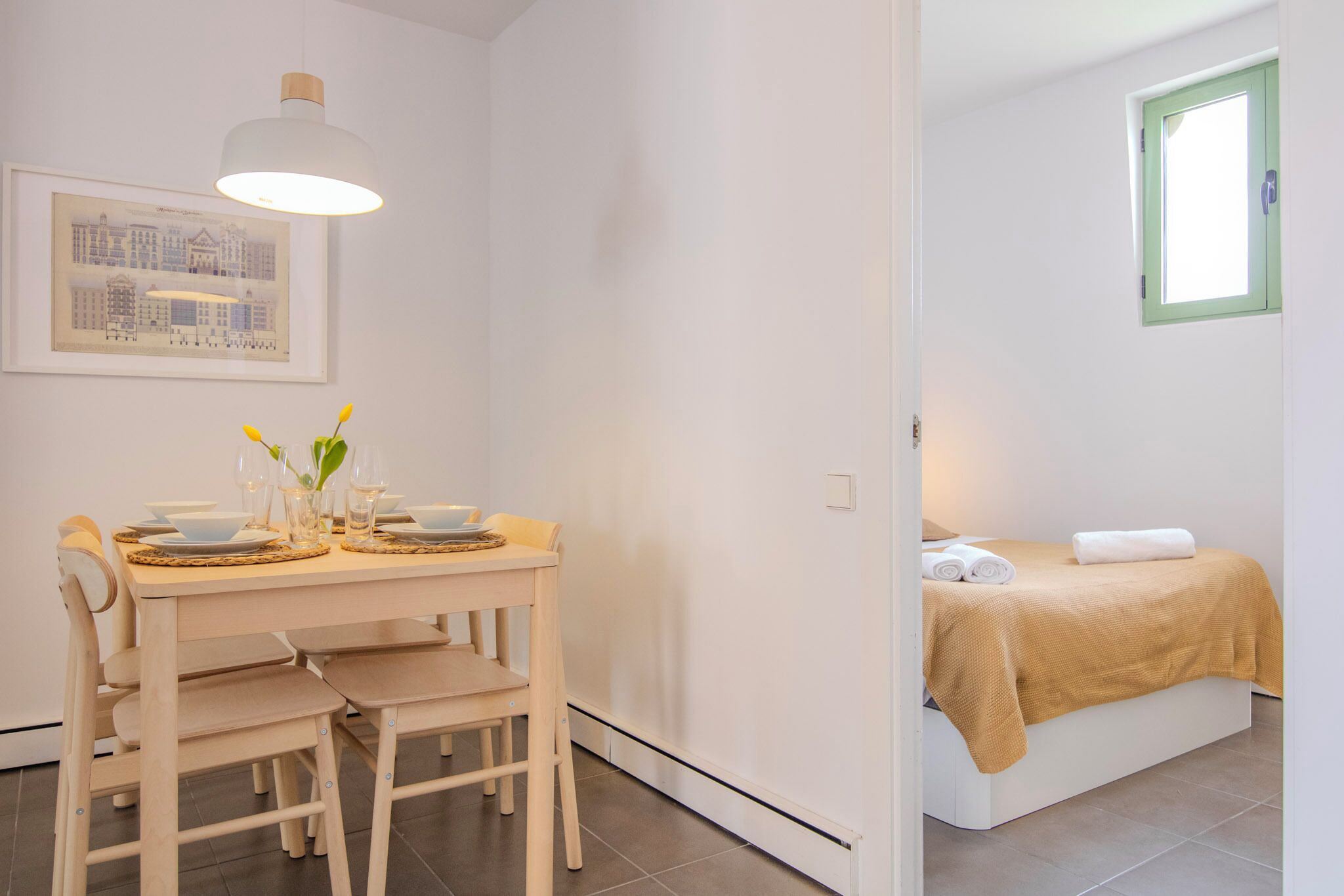 A room with a dining table set for two, a bed with a green window, and a framed cityscape on the wall.