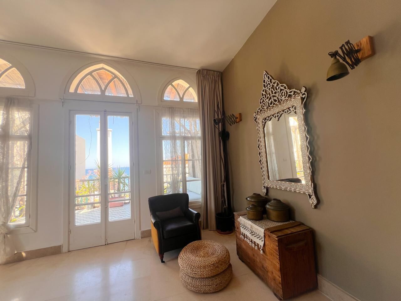Una habitación amplia con un ventanal grande, un sillón negro, un otoman tejido y un mueble de madera con objetos decorativos.