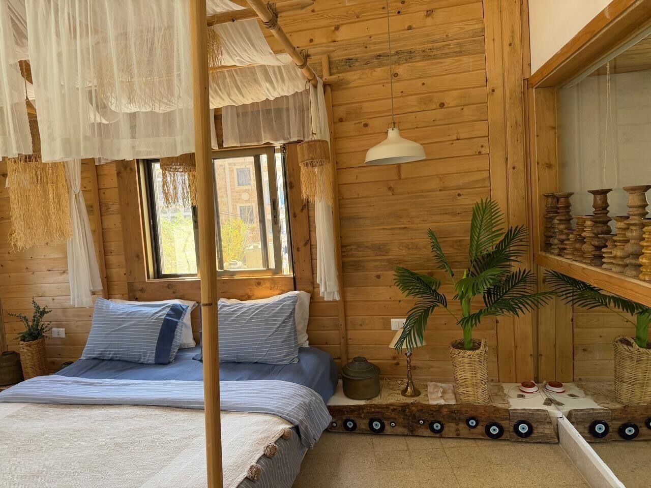 A wooden cabin bedroom with a bed, hanging curtains, a lamp, and potted plants.