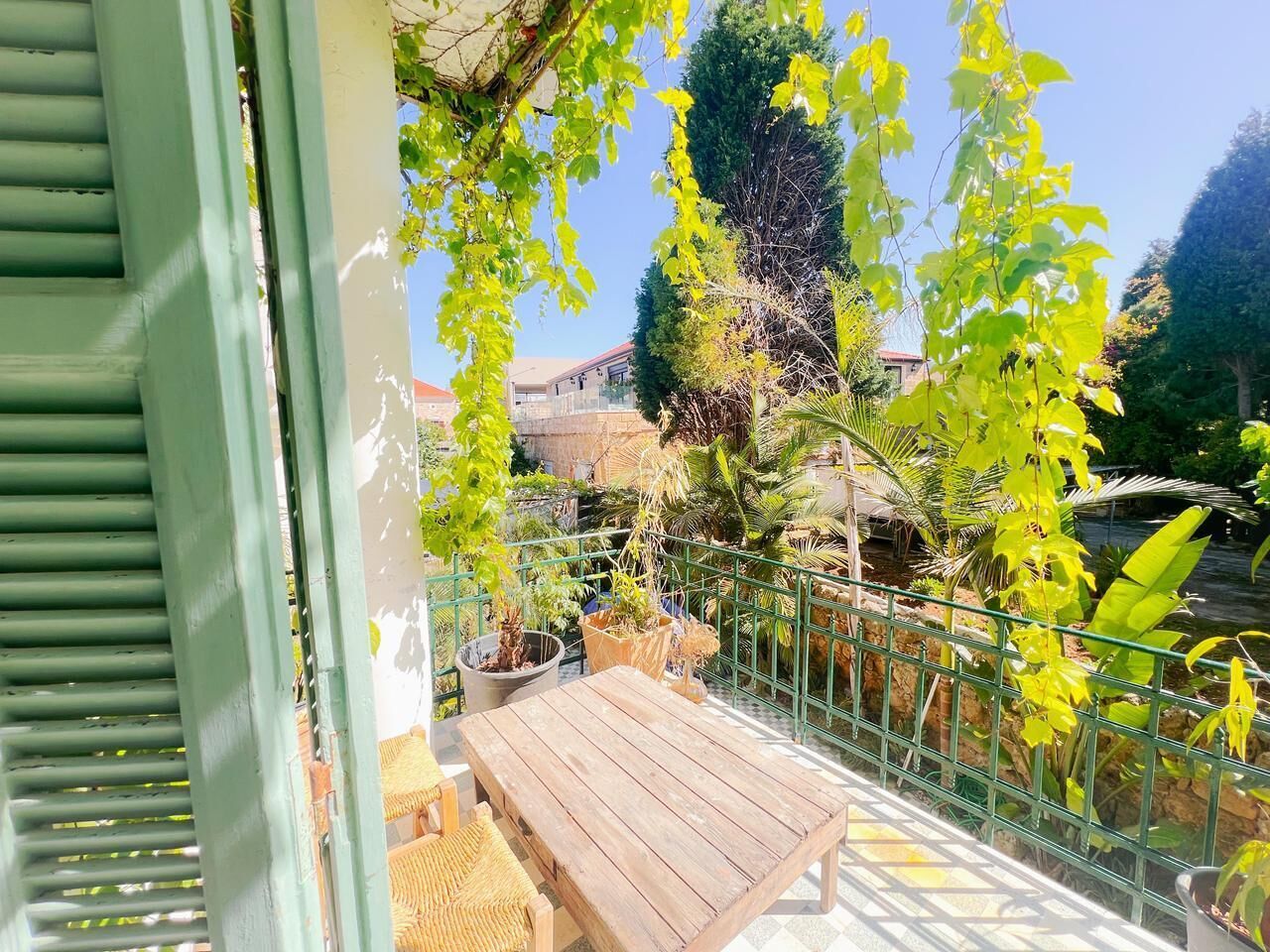 Une table et des chaises en bois sur un balcon doté d’une rambarde en métal, entourés de plantes en pot, avec en arrière-plan la vue d’un immeuble.