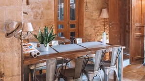 Ein rustikaler Raum mit Steinwänden, einem Holztisch und Metallstühlen. Auf dem Tisch stehen ein Blumentopf und ein Hut.
