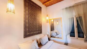 A bedroom with a large bed, a hanging light fixture, a patterned tapestry, a mirror, and a window with curtains.