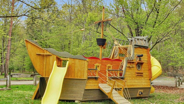 A wooden pirate ship play structure with a slide and climbing area, surrounded by trees. - Full Hook up 30 Amp Site W Up to 26Ft (Wellsboro)