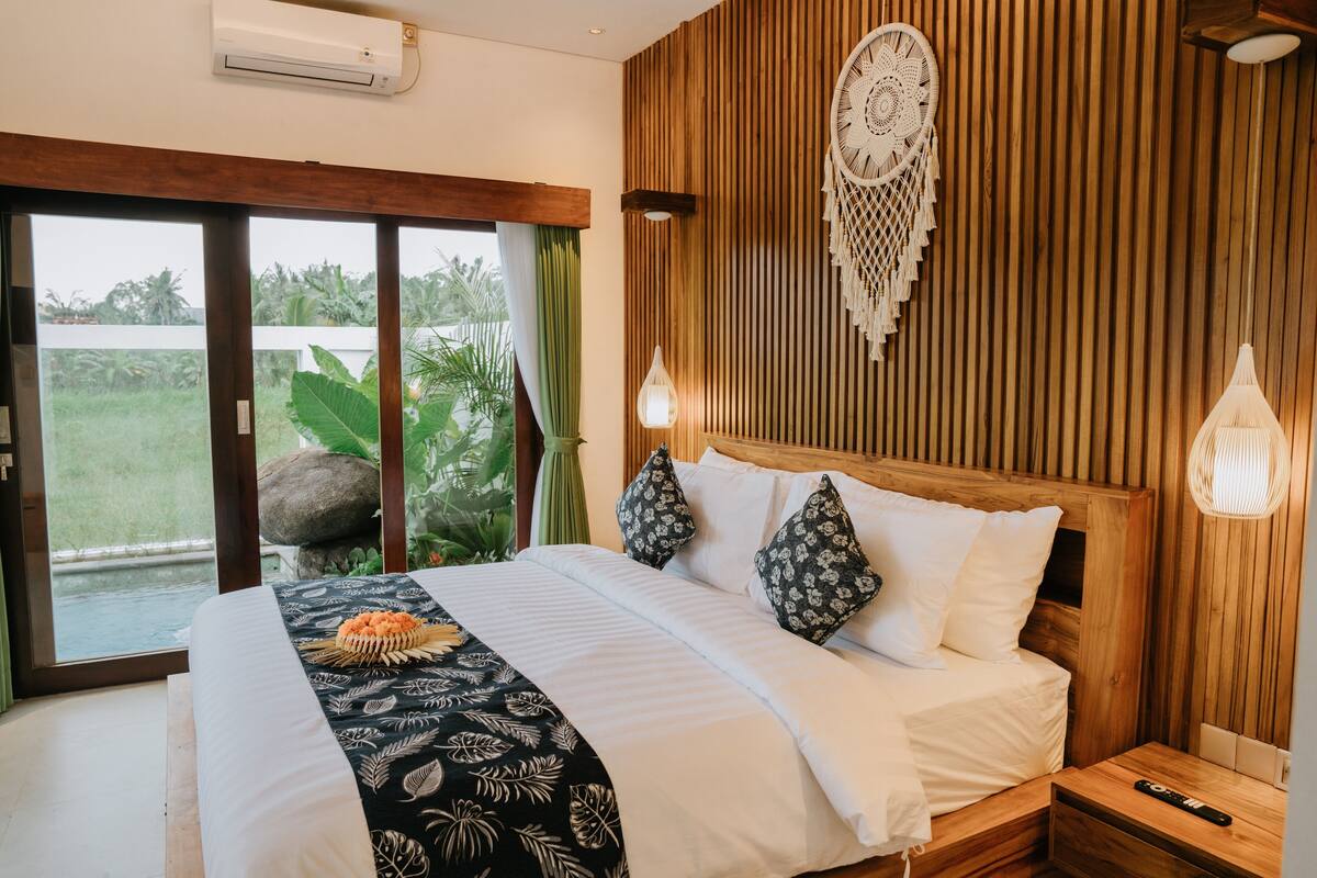 A bedroom with a bed, wooden headboard, bedside lamps, and a view of greenery outside.