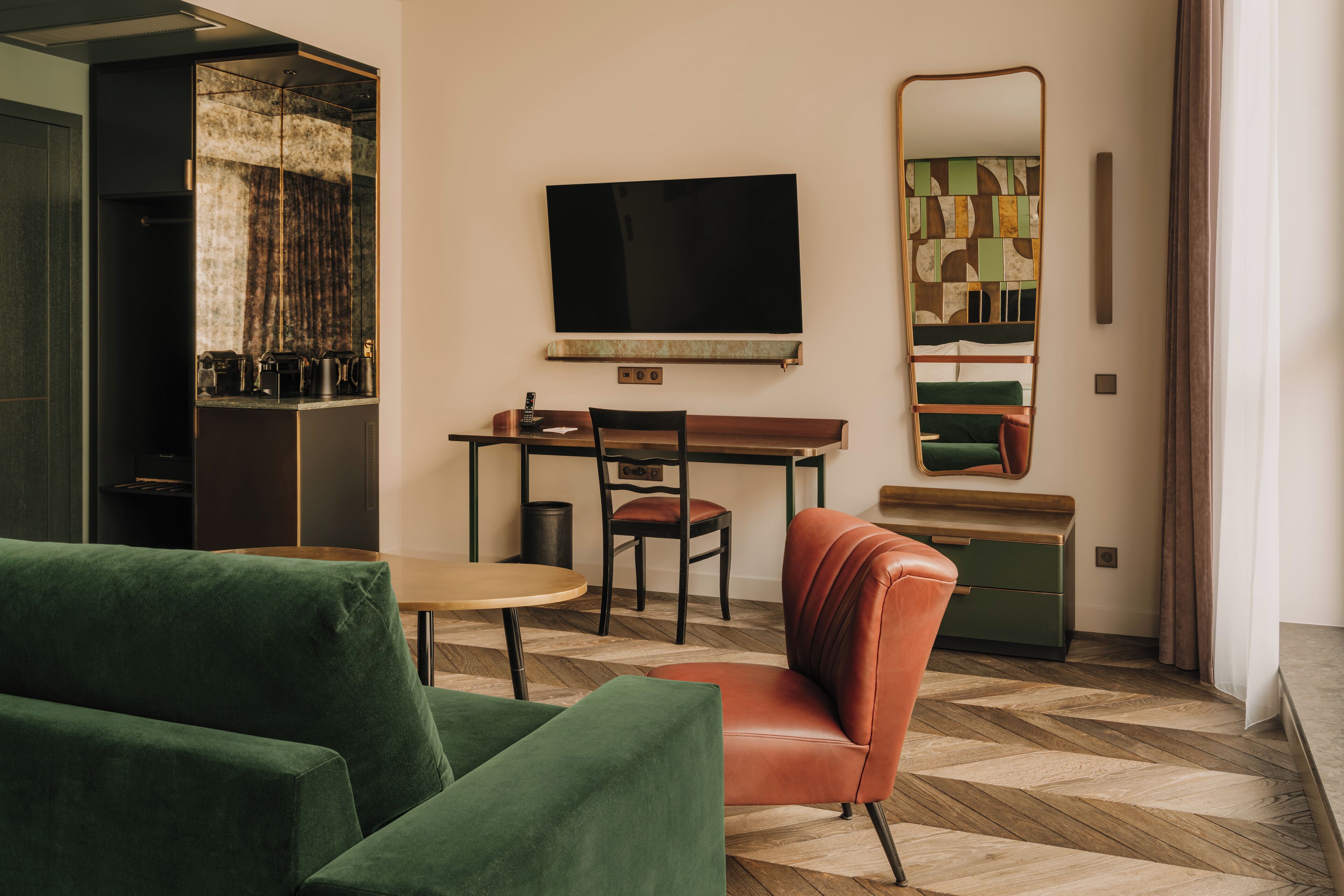 A modern hotel room with a large mirror, a flat-screen TV, a wooden desk, a green sofa, and a herringbone-patterned floor.