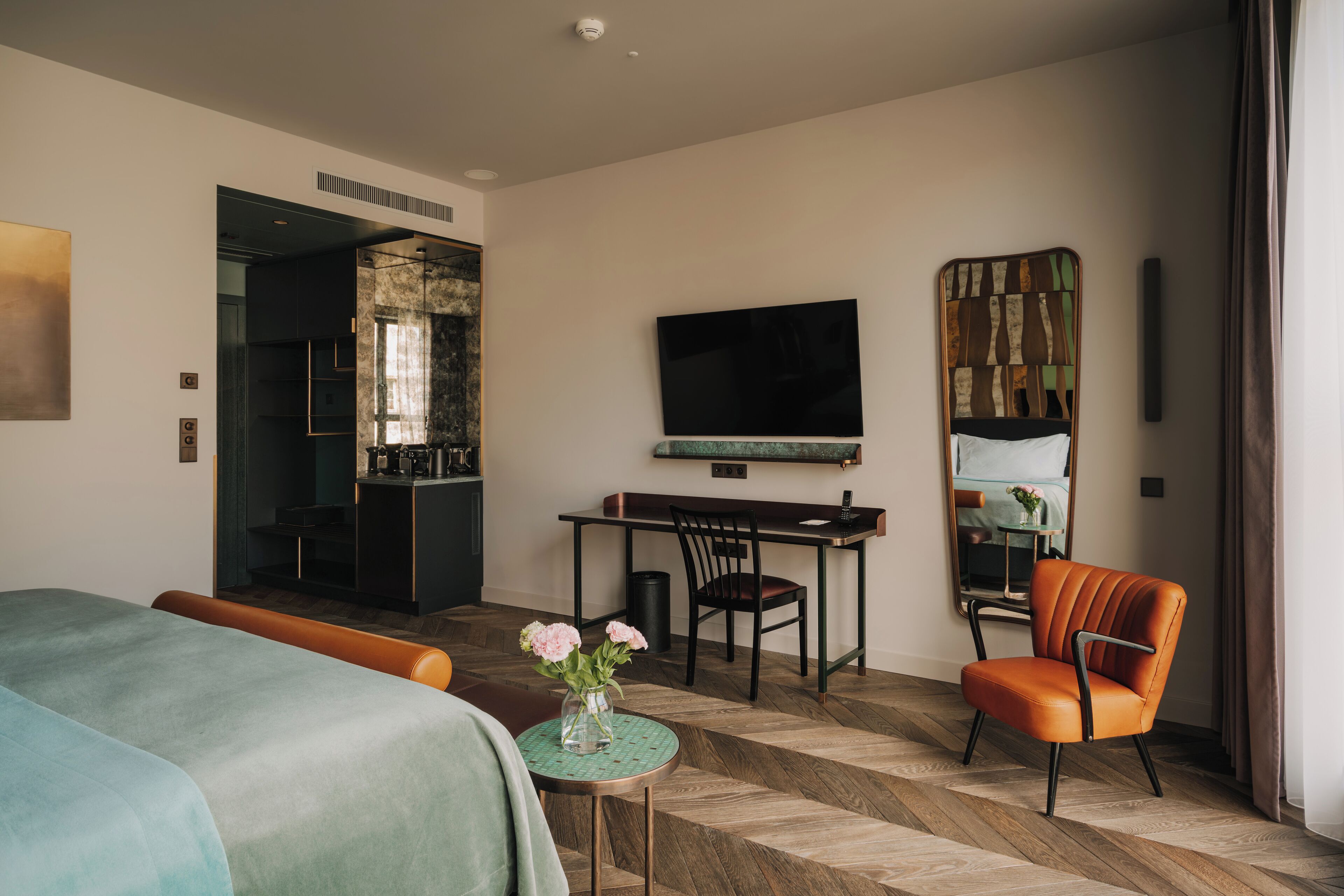 A hotel room with a bed, a desk, a chair, a TV, and a small table with flowers.