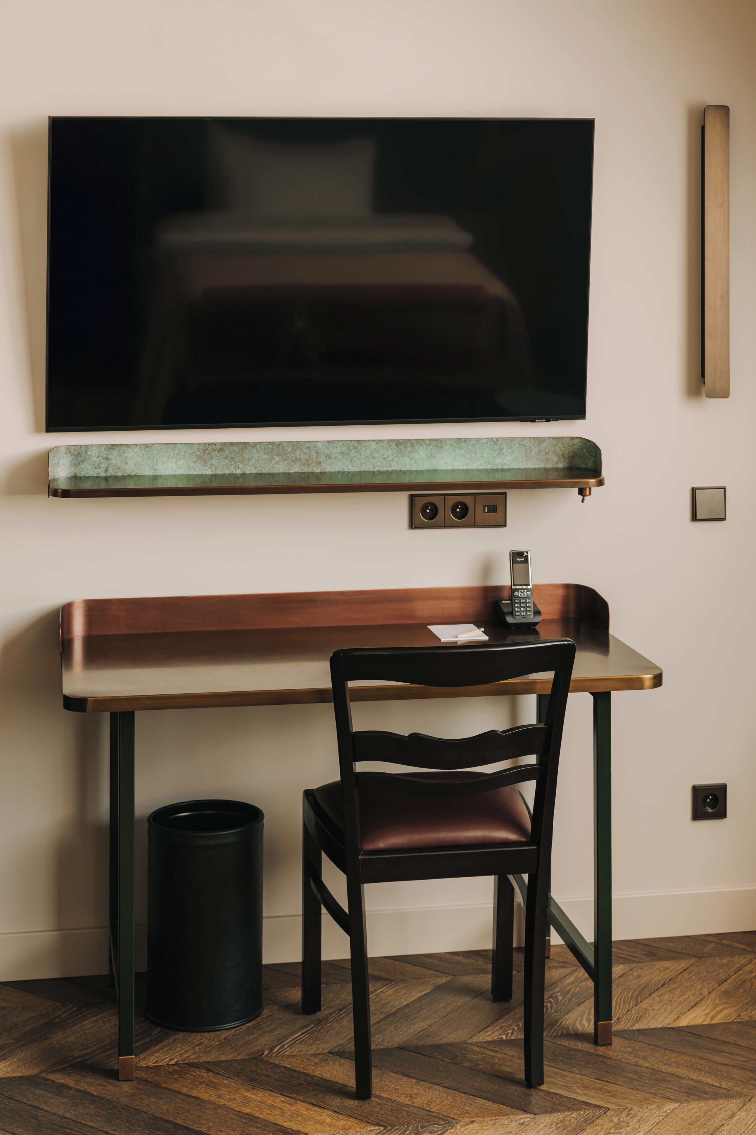 A modern hotel room with a wooden desk, a black chair, a television mounted on the wall, and a trash can.