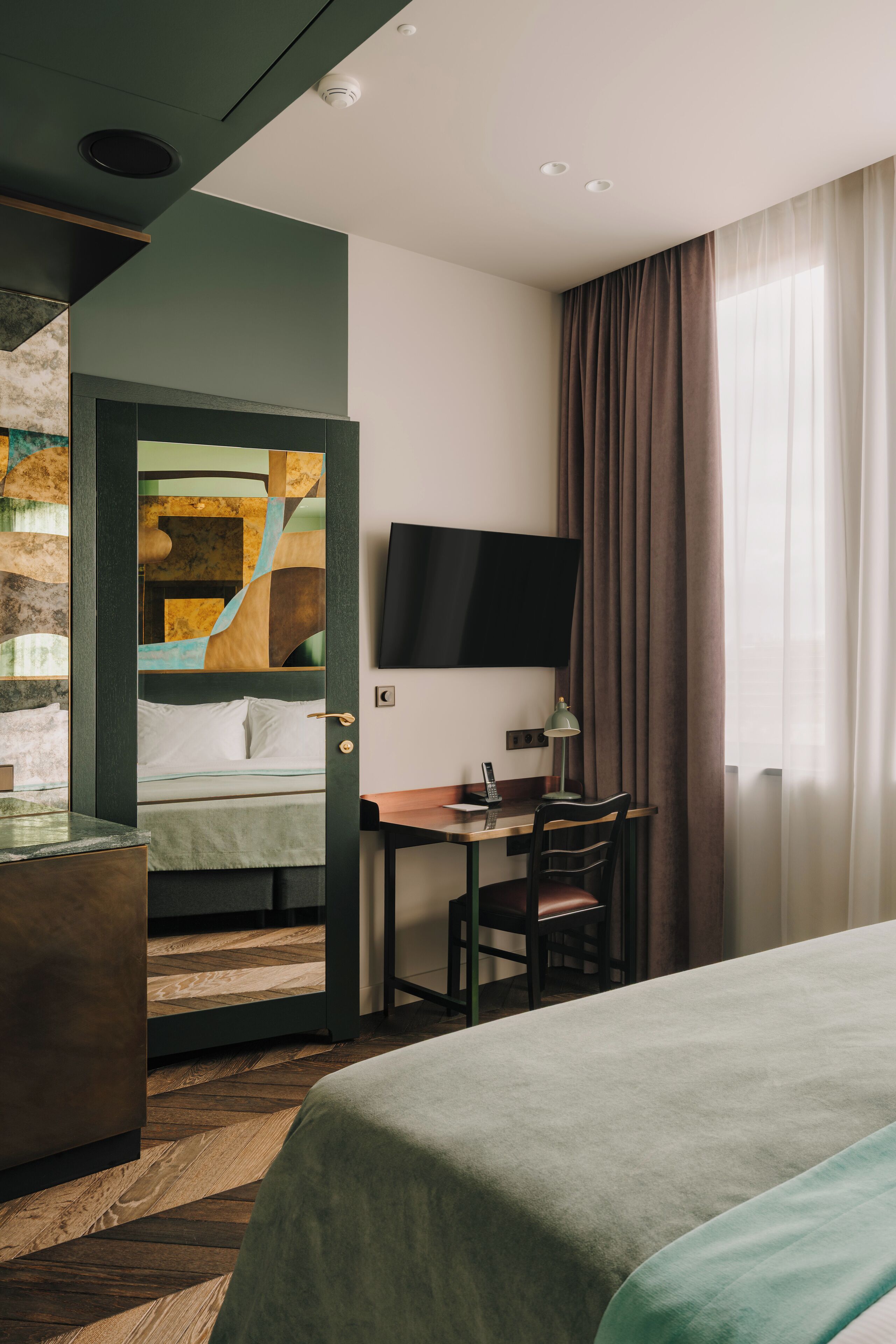 A hotel room with a bed, a desk with a chair, a television on the wall, and a window with curtains.