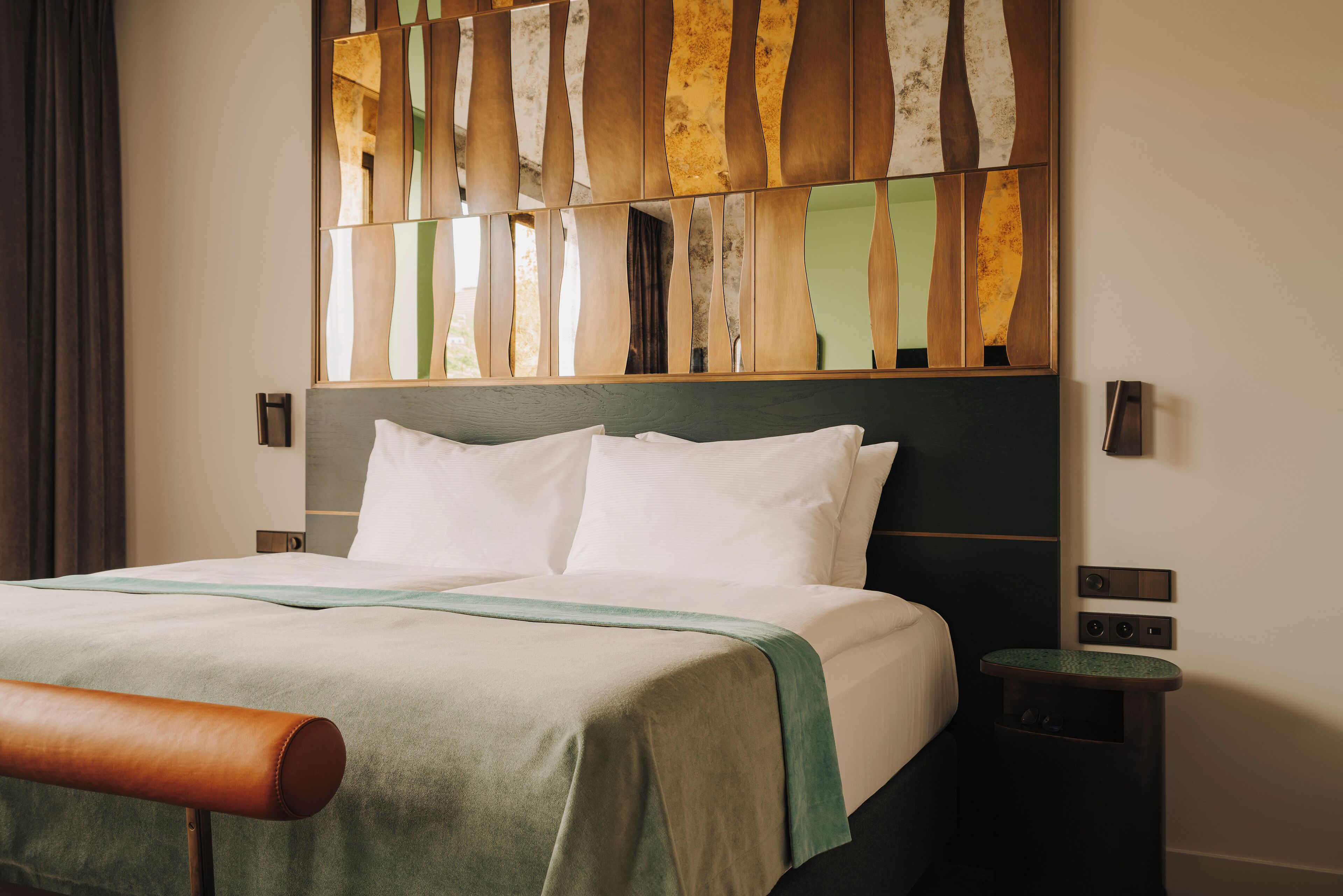 A neatly made bed with white linens, a green and brown bedspread, and a wooden headboard with a decorative panel.
