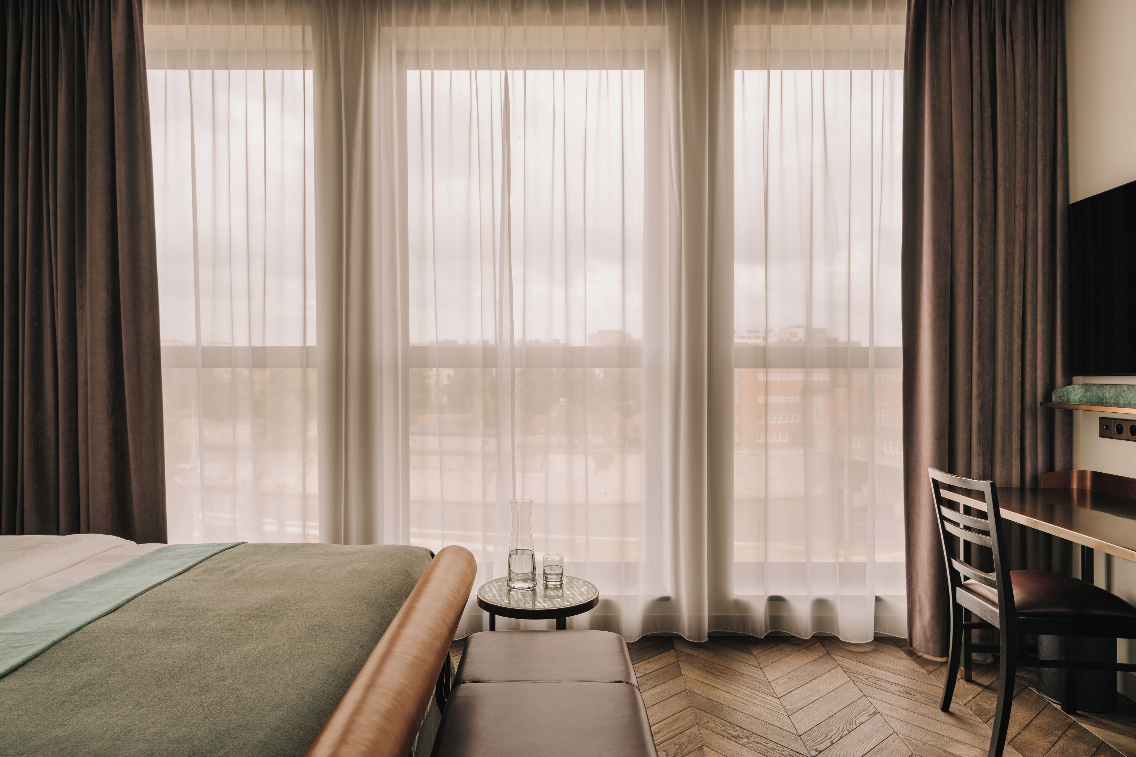 A hotel room with a bed, a small table with glasses, a chair, and a view through sheer curtains.